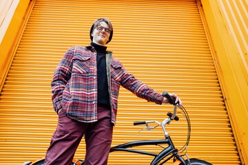 Bottom view Young stylish hipster man standing with a cruiser city Bicycle near orange Wall....