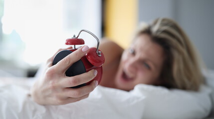 Screaming woman in bed with a red alarm clock in her hand, close-up, blurry. Oversleep for work,...