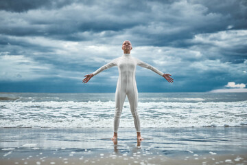 Happy hairless girl with alopecia in white futuristic suit stands with spread arms on beach bathed...