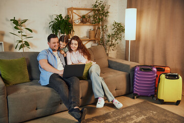 Fototapeta premium Choosing destination via Internet. Happy family, mother, father and daughter sitting at home, looking on laptop and planning upcoming trip. Concept of vacation, traveling, booking services