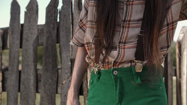 Young stylish slim guy wearing rustic country style jeans with suspenders and plaid shirt poses by a wooden fence 