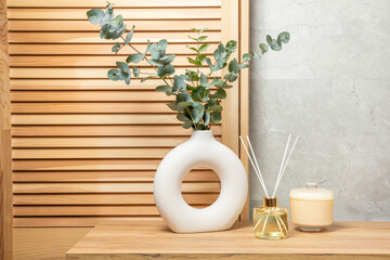 Green eucalyptus leaves in a vase stand on a chest of drawers against the background of a wall. Aromatherapy.Beautiful eucalyptus bouquet.Minimalist interior with flowers, candles and aroma diffuser. © Avocado_studio