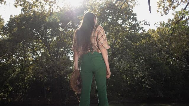 Traveler with straight long hair wearing a checkered shirt stands by a river in the woods in bright sunny weather 