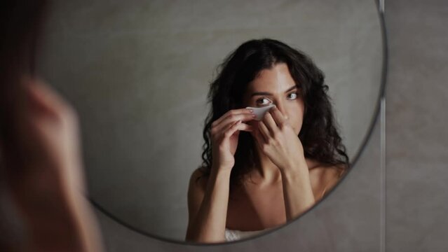 Medium close-up selective focus shot of beautiful young Caucasian or Hispanic woman, wearing bath towel around chest, applying moisturizing under eye patches then looking at herself in mirror