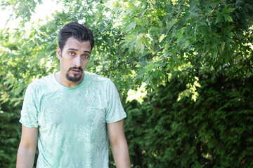 A wet and upset man stands in the park. The guy was caught in a sudden rain in broad daylight