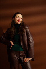 Fashionable happy smiling woman wearing faux leather suit blazer, pants, green turtleneck, posing on brown background. Studio fashion portrait