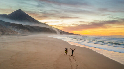 Aerial view of two people waving while walking on Cofete beach at the sunset. Above view of the large beach of Cofete and monolith Roque del Moro in the south island of Fuerteventura, Canary Islands.