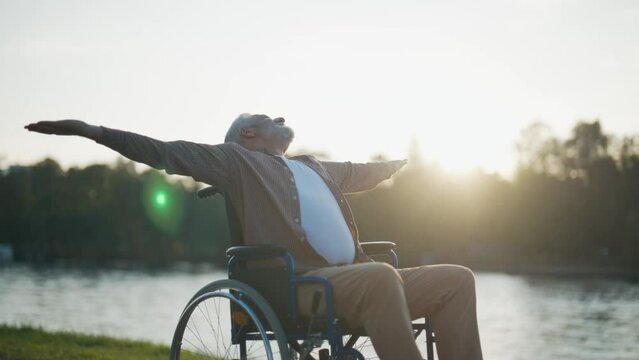 Senior man in wheelchair enjoys freedom. Male with special need spreading arms on sides resting breathing fresh air walking in city park near pond. Living with physical disability in old age concept.