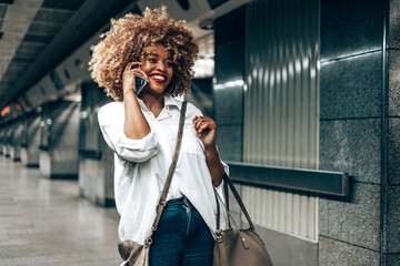 Beautiful fashionable black woman standing at a subway train station. She is happy and talking to someone on her smart phone. Public transportation and urban life concept.
