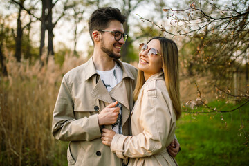 A man looks at a woman with love and tenderness. Close shot of a beautiful young couple in glasses and beige trench coats posing against the backdrop of a spring forest park.