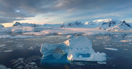 Sunset Antarctic iceberg floating in icy ocean. Snow-capped mountains in background. Fly over the...