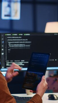Software developer programming in home office, crosschecking lines of code between tablet and PC screen. IT admin holding portable device, using it as reference while coding on computer, camera A