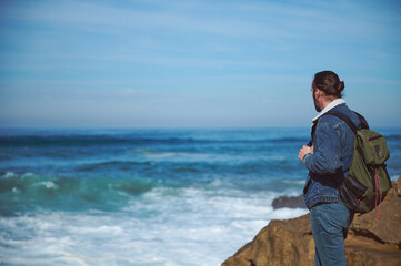 Caucasian young bearded traveler with backpack, watching the horizon standing on a cliff by the see...