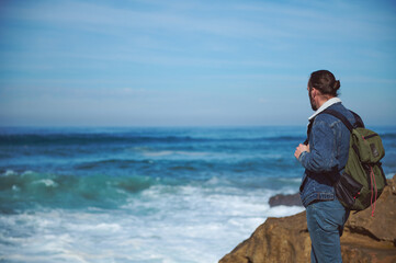 Caucasian young bearded traveler with backpack, watching the horizon standing on a cliff by the see in a sunny windy day
