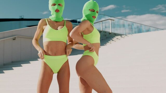 Two beautiful sexy women in green underwear. Models wearing bandit balaclava mask. Hot seductive female in nice lingerie posing in the street at sunny day, blue sky. Crime and violence. Perfect body 