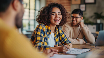 Cheerful professionals in casual wear engage in a lively discussion during a collaborative meeting, with one person taking notes and others interacting around a wooden table with laptops.