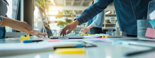 close-up view of a person's hands working on a laptop with other individuals in the background, surrounded by colorful sticky notes, papers, and a collaborative work environment.