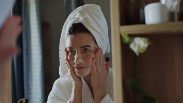 Close-up over-shoulder shot of Caucasian or Hispanic woman in bathrobe and towel turban, standing in front of mirror and massaging peeling face mask over clean skin with gentle movements
