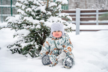 A child dressed in winter clothes smiles against the background of a snow-covered Christmas tree, it is snowing