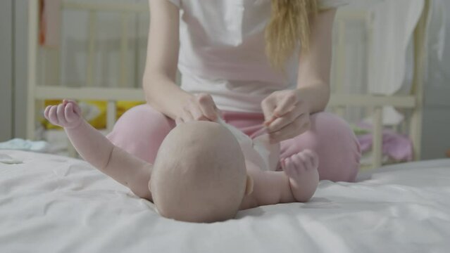 Infant lies on back during diaper change, mother hands show, nursery room setting. Clean, simple newborn care scene, baby relax on white bedding, curious child leg lift, focus on hygiene and comfort.