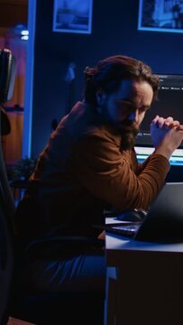 Software programmer putting headphones on while developing code, making time pass faster during remote job shift. IT professional doing multitasking in apartment office while listening music, camera B