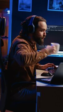 Programmer does multitasking to finish developing PC code, drinks coffee, listens music. IT admin wearing headphones, enjoying hot beverage, solving database errors on computer and laptop, camera B