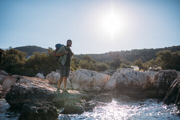 A male tourist with a large backpack and hiking gear on a rocky seashore.