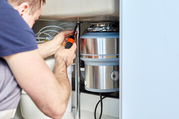 A plumber with gloved hands installs a household waste shredder for the kitchen sink. A technician installs or repairs a household waste shredder in the kitchen.