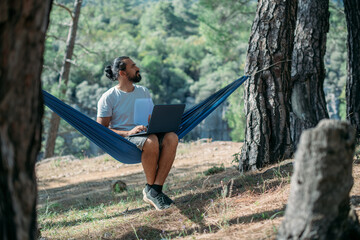 A young man is working on a laptop while lying in a hammock in the mountains.