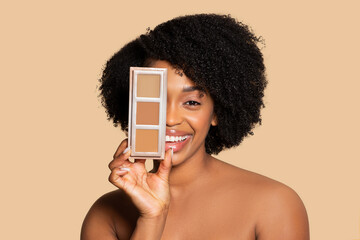 Happy young black woman holding concealer palette near face