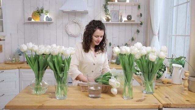 Beautiful caucasian woman puts chic bouquet of fresh white tulips in vase on the kitchen. Girl surrounded by flowers. Bunch of first spring flowers. Concept for floristic business or flower delivery