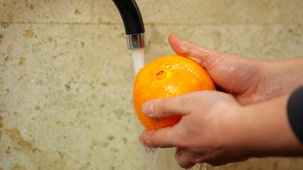 Male hands wash an orange under the tap in the kitchen. close-up.  Hygiene, microbes on fruit....