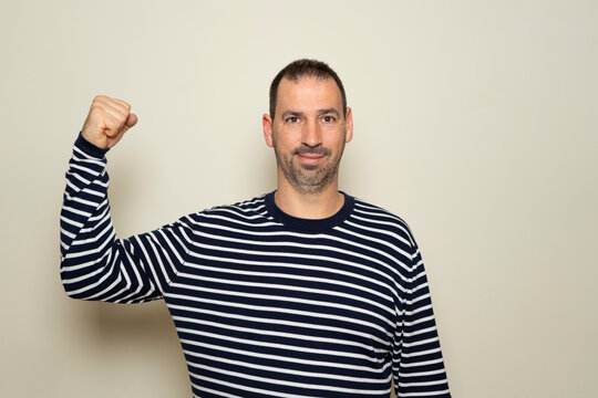 Hispanic man with a beard of 40 years old showing biceps believing that his training has paid off, the poor deluded man does not realize his terrible physical condition. Isolated beige background.