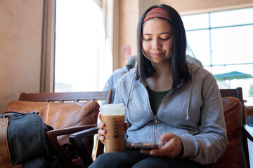 Young student woman using her mobile phone and drinking a cold brew coffee before classes at...
