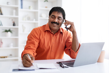 Obraz premium Smiling mature indian man working on laptop, have phone conversation