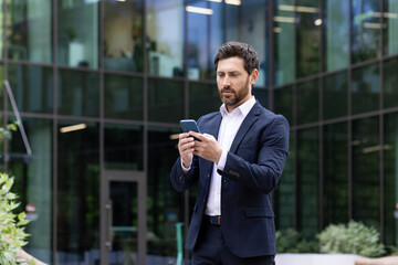 Confident bearded businessman using smartphone in front of modern office