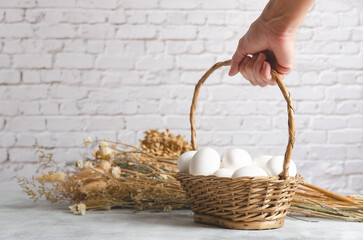 Female hand holding a basket with white chicken eggs on a light background with dried flowers....