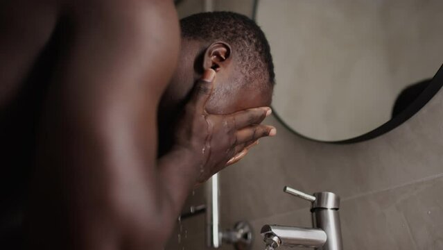 Close-up side shot of young black man with short hair and bare chest bending over bathroom sink, taking handful of running water, washing and rubbing sweaty face, then standing up and shaking hands