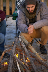 Campfire, wood and men by beach for travel on vacation, adventure or holiday camping. Nature, outdoor and young male people sitting on chair in sand with flame for heat on weekend trip in winter.