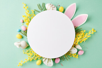 Easter greeting: spring at heart. Top view photo of Easter eggs, mimosa, bunny ears on a teal background with blank circle for writing festive greetings or advertising copy