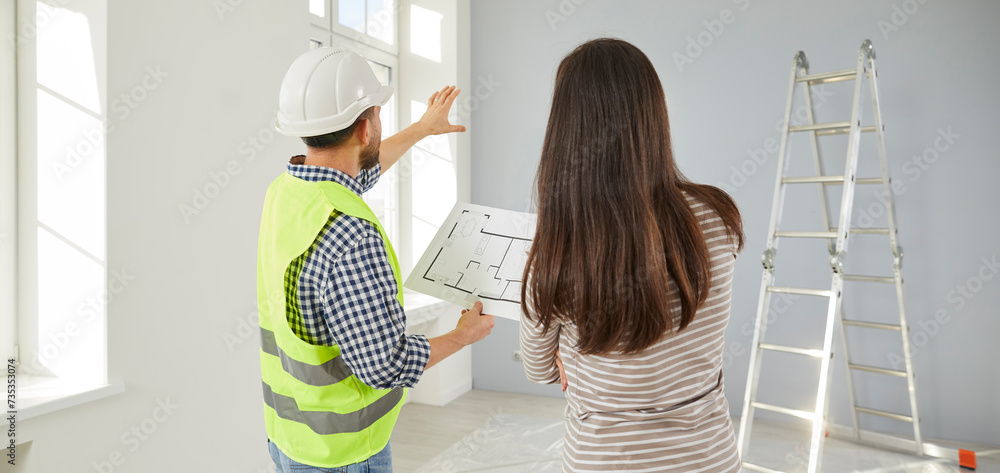 Wall mural Architect or builder in uniform hard hat standing in living room at home, holding blueprint construction plan, and talking to young woman about house conversion, replanning design and wall demolition