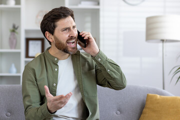A bearded middle-aged man is engaged in an animated phone conversation, sitting alone on a sofa in...