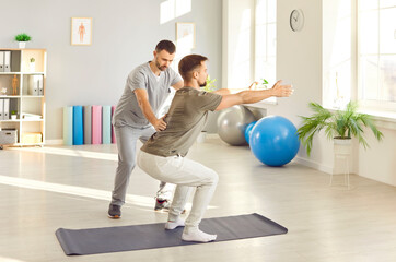 Male nurse, caregiver or physiotherapist helping young man in doing sport exercise on yoga mat in...