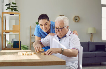 Development of senior people skills. Friendly, caring nurse helps elderly man correctly combine and assemble wooden puzzle pieces. Yyoung female caregiver and male pensioner in nursing home.