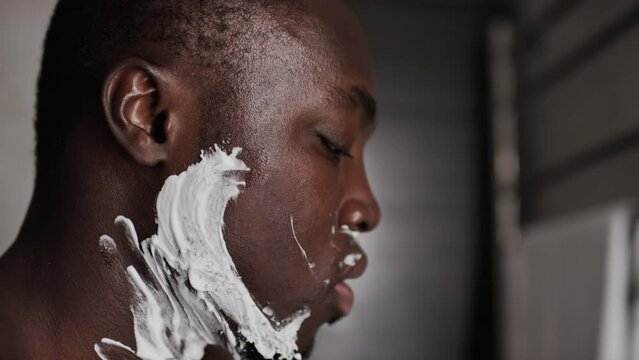 Close-up side shot of young African American guy with cropped hair shaving sideburns and beard over white foam with disposable razor at home