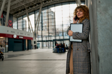 Amidst the airport buzz, a content Caucasian woman leans against a wall, talking on the phone and cradling a laptop, a harmonious blend of travel, communication, and technology.