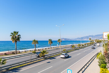 Obraz premium Highway Road with Cars Moving Next to a Blue Sea and Sandy Beach Under The Clear Sky in a Hot Sunny Summer day from a Pedestrian Stone Bridge