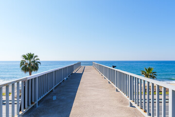 Empty Stone Pedestrian Crosswalk Bridge Over a Highway with a Blue sea and Palm Trees Behind it Under the Clear Sky in a Hot Summer Day