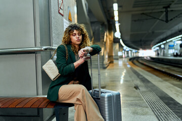A weary businesswoman with curly hair sits, patiently awaiting her train at the station.