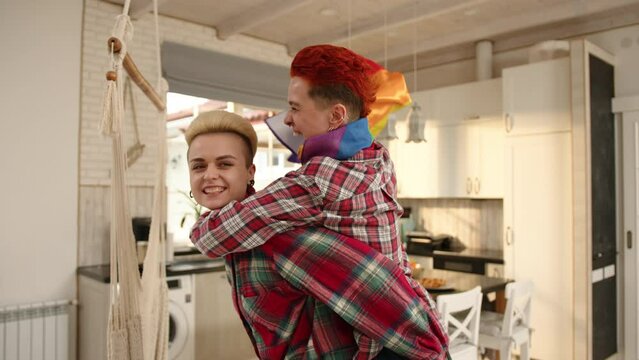 Two lesbians share a playful moment at home, one riding piggyback on the other, gleefully waving an LGBT flag, a spontaneous expression of their joy and pride. Camera 8K RAW. 